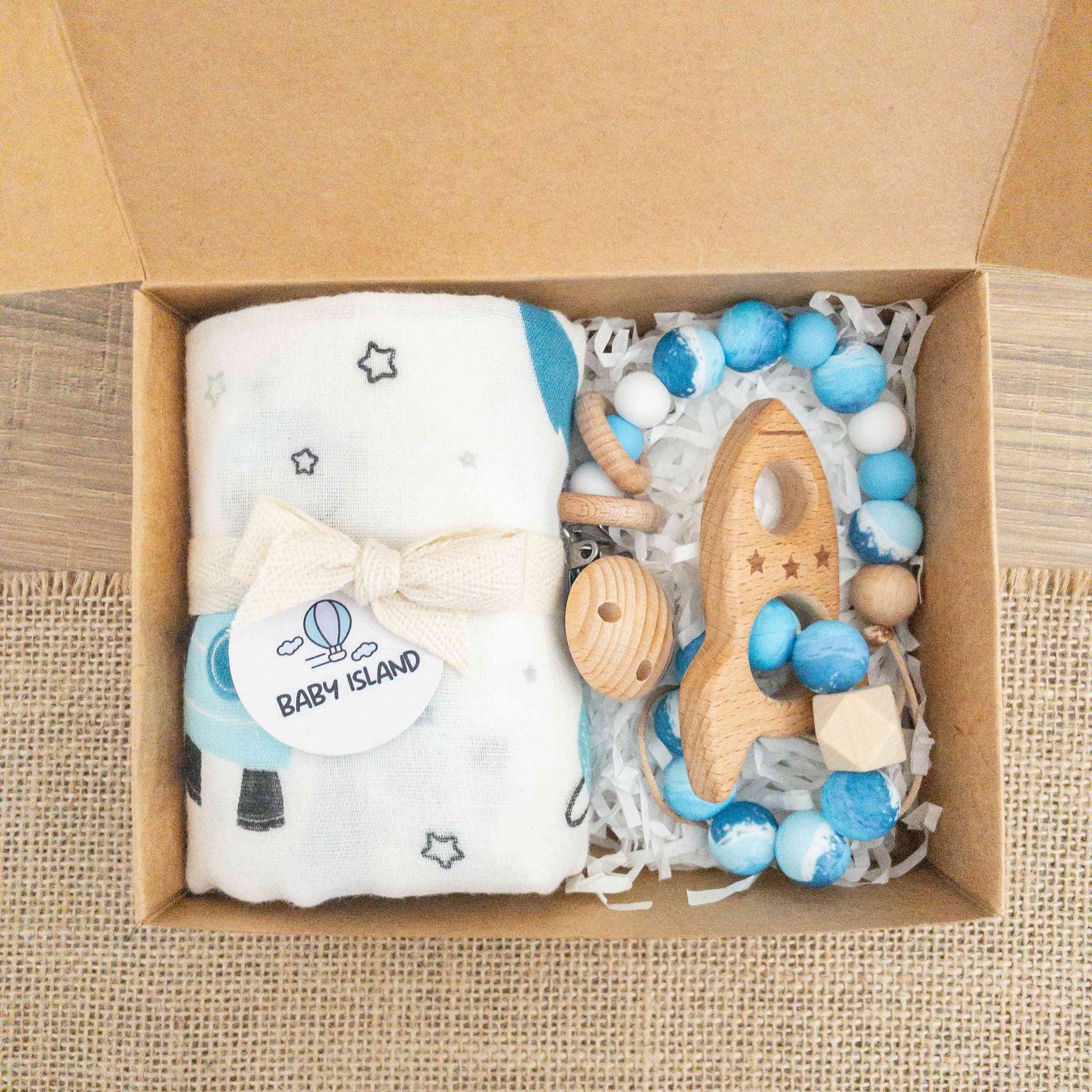 scatola con set regalo neonato con mussola a tema spazio, pianeti e razzo spaziale. catenlla ciuccio clip con perline nei colori azzurro bianco blu e massaggiagengive in legno a forma di razzo