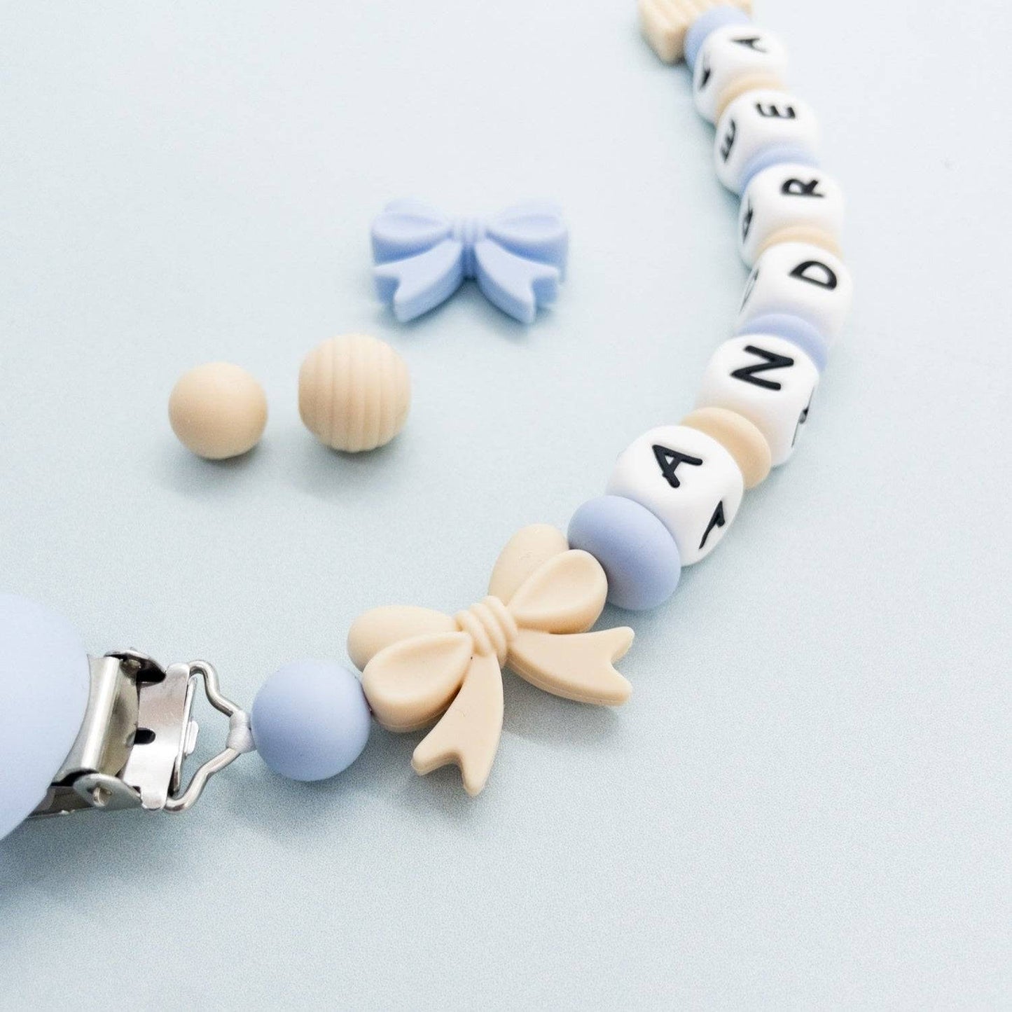 Catenina porta ciuccio personalizzata per neonati in silicone con perline beige e azzurre, decorata con fiocchetti, ideale per la dentizione e per mantenere il ciuccio a portata di mano in sicurezza