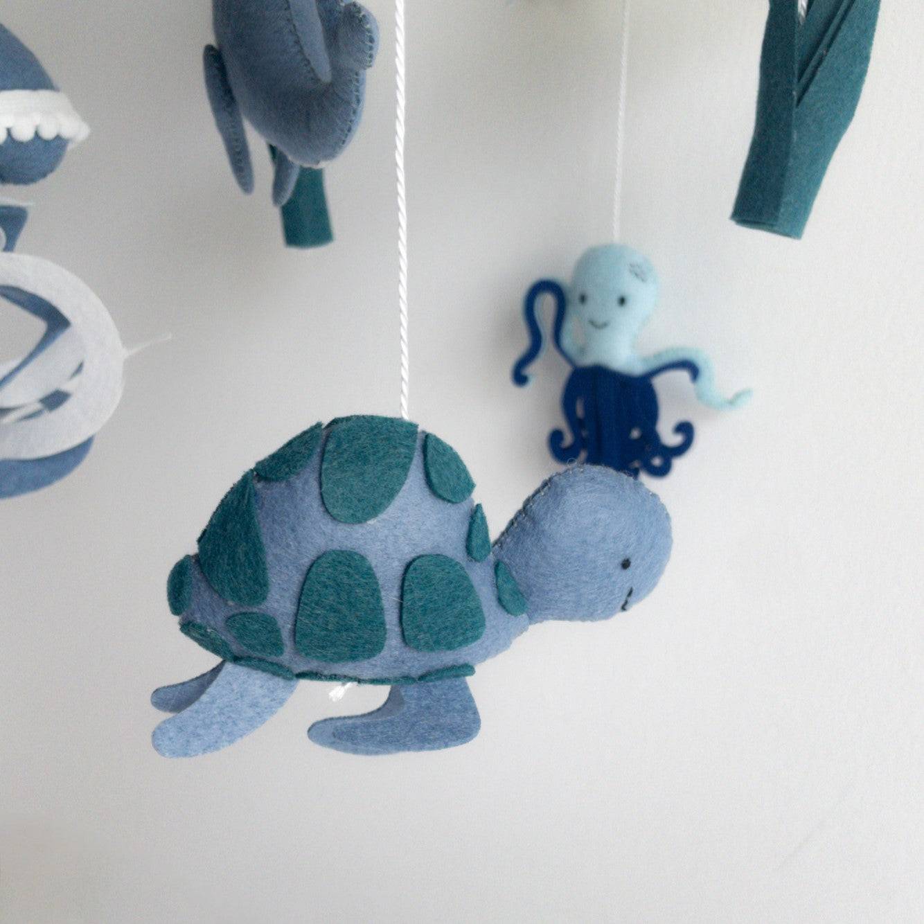Elemento decorativo di una giostrina culla neonato a tema mare con targaruga, polpo di stoffa blu e celeste sospeso, accanto a nuvole e alghe di feltro verde