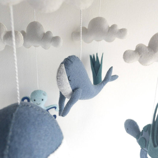 Giostrina per culla a tema marino con una balena di feltro blu e bianco e una tartaruga verde e blu, appese a fili sotto nuvole di stoffa