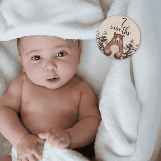 Foto di un neonato sorridente sdraiato su una coperta a tema foresta, affiancato da una milestone card in legno con la scritta “7 months old”. Perfetta per immortalare il settimo mese del bambino, questa immagine è ideale per genitori che desiderano documentare la crescita del proprio piccolo. Parte del set regalo Woodland Adventures, include accessori a tema animali della foresta