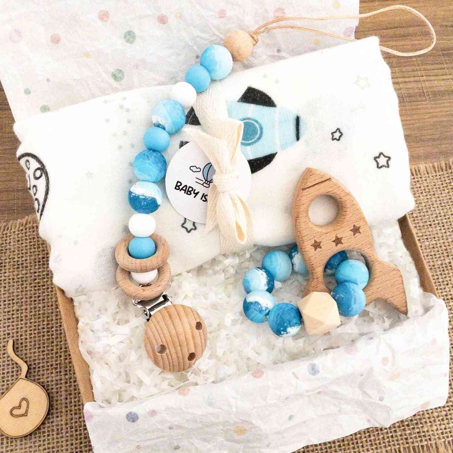 scatola con set regalo neonato con mussola a tema spazio, pianeti e razzo spaziale. catenlla ciuccio clip con perline nei colori azzurro bianco blu e massaggiagengive in legno a forma di razzo