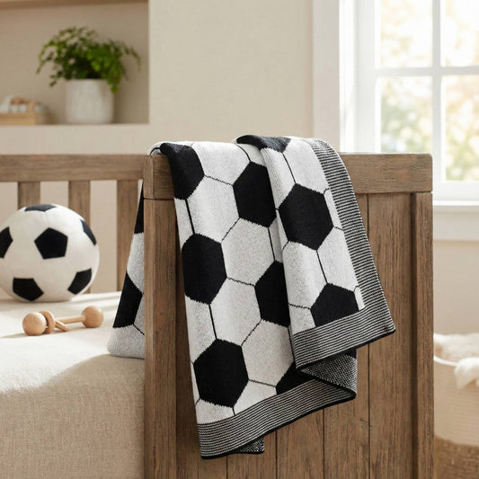 Coperta per neonato 100% cotone con motivo calcio in bianco e nero, appoggiata su lettino in legno, perfetta come regalo a tema sportivo.