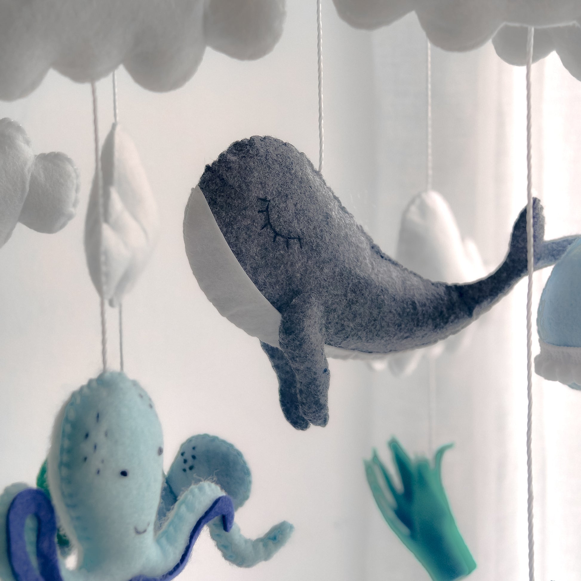 Accessorio di design per nursery a tema sottomarino. Giostrina pendente che aggiunge un tocco di colore blu e verde alla stanza del bambino. Elementi fluttuanti tra nuvole bianche che creano un ambiente sereno e sognante sopra il fasciatoio o la culla