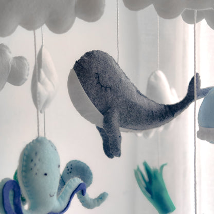 Accessorio di design per nursery a tema sottomarino. Giostrina pendente che aggiunge un tocco di colore blu e verde alla stanza del bambino. Elementi fluttuanti tra nuvole bianche che creano un ambiente sereno e sognante sopra il fasciatoio o la culla