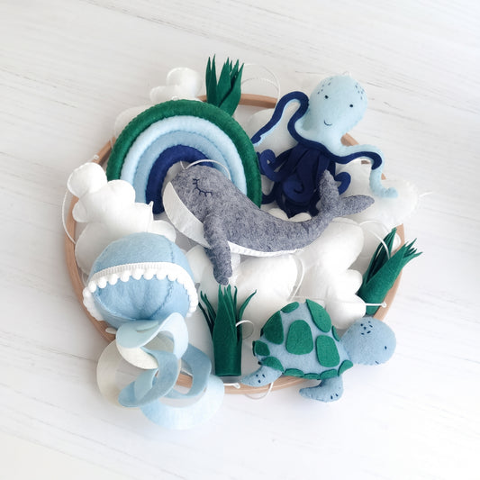 Giostrina culla a tema mare per lettino con palette colori rilassante blu, verde salvia e grigio. Figure marine in feltro imbottito che fluttuano sotto un cerchio in legno naturale. Include alghe marine e nuvole soffici, ideale per arredamento nursery moderno o stile nordico scandinavo per bambini