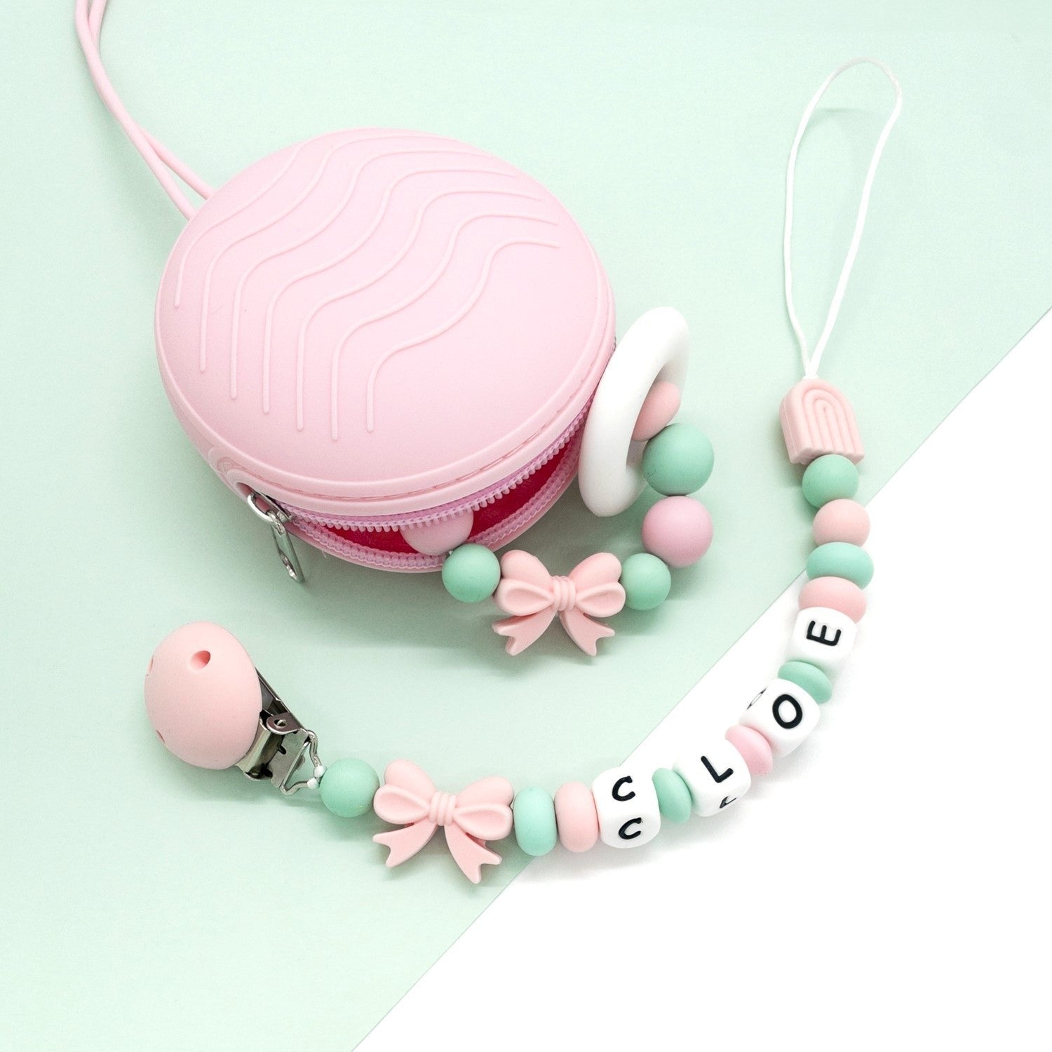 Set di accessori per neonati in silicone con custodia rosa, massaggiagengive ad anello e catenina porta ciuccio personalizzata con nome 'Cloe', in tonalità pastello rosa e verde menta su sfondo minimal verde e bianco