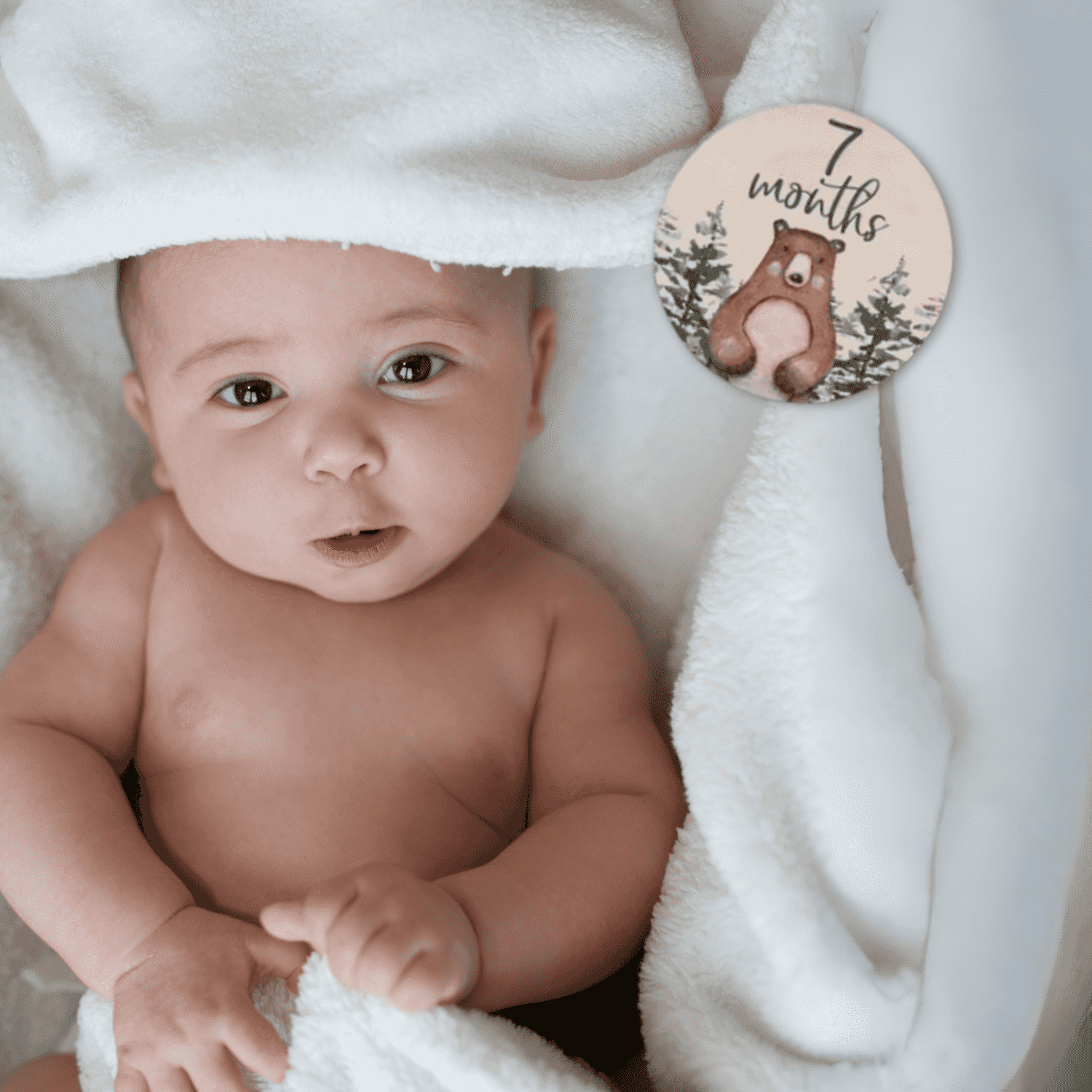 Foto di un neonato sorridente sdraiato su una coperta a tema foresta, affiancato da una milestone card in legno con la scritta “7 months old”. Perfetta per immortalare il settimo mese del bambino, questa immagine è ideale per genitori che desiderano documentare la crescita del proprio piccolo. Parte del set regalo Woodland Adventures, include accessori a tema animali della foresta