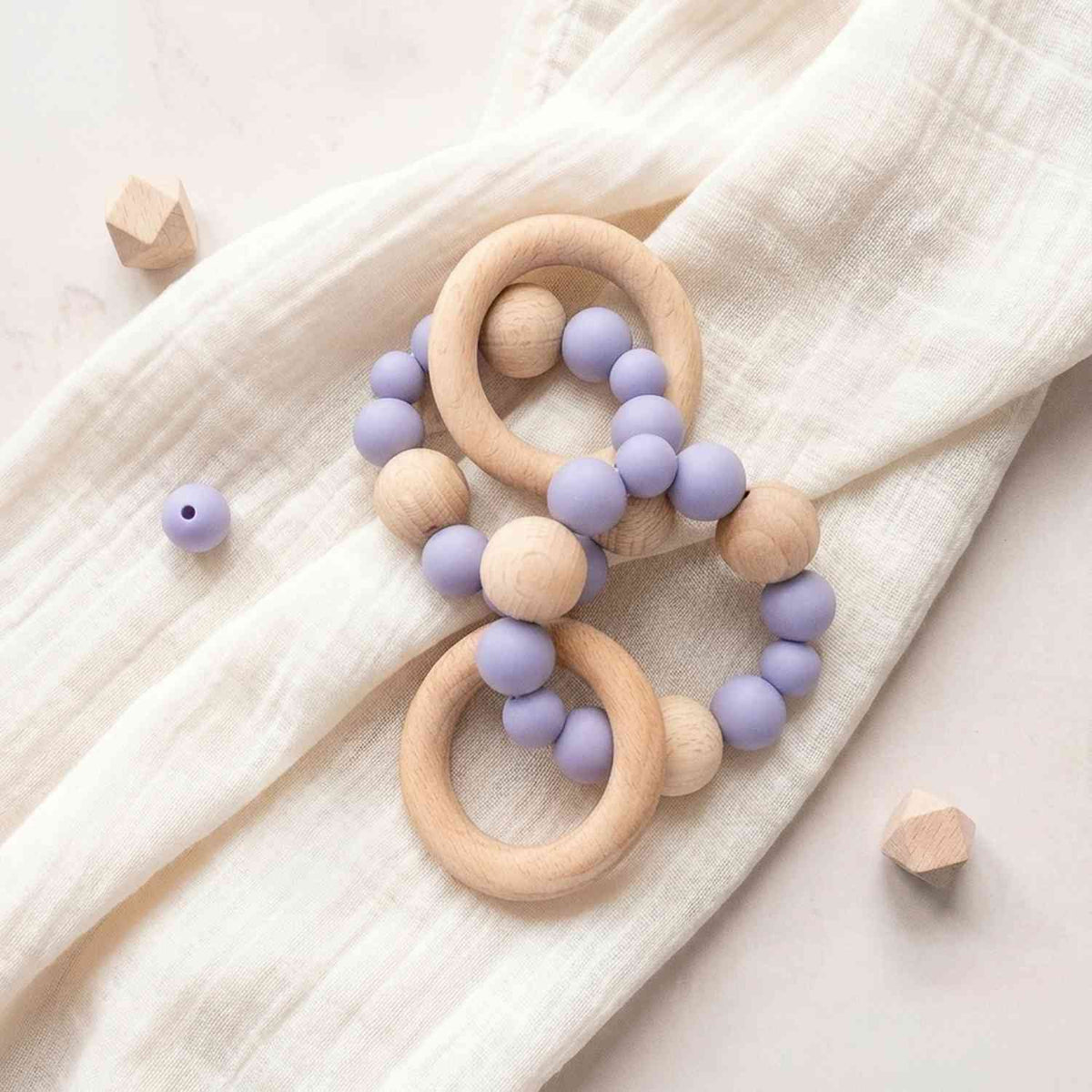 Dettaglio di ciuccio in legno naturale con palline in silicone lilla e stelle in legno, su tessuto lilla morbido, confezione regalo elegante