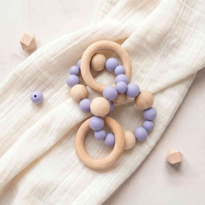 Dettaglio di ciuccio in legno naturale con palline in silicone lilla e stelle in legno, su tessuto lilla morbido, confezione regalo elegante