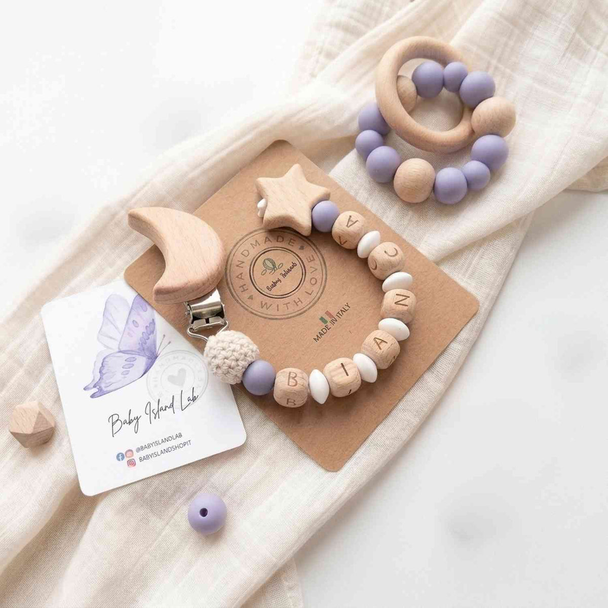 Dettaglio di ciuccio in legno naturale con palline in silicone lilla e stelle in legno, perfetto come regalo neonato, su tessuto lilla